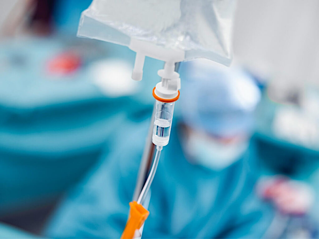 Medicine Drips from Plastic Tubing and Plastic IV bag That have been Sterilized Using Ethylene Oxide (EtO) Gas. A Nurse Wearing Blue Full Surgical Garb and a Patient are in the background