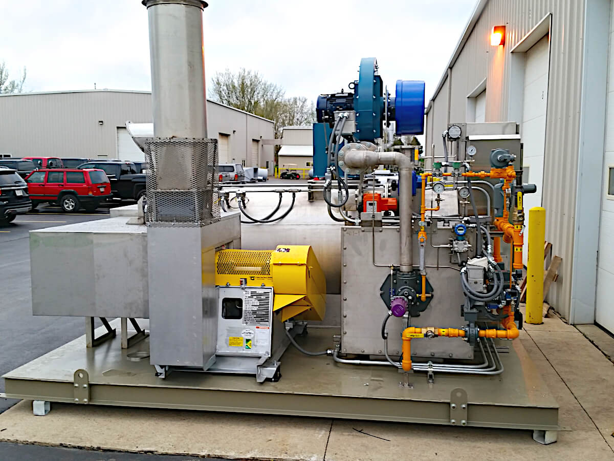 A skid-mounted silver Electric Catalytic Oxidizer waits in a parking lot for shipping to an automotive facility. It has bright yellow components and a blue fan. This small ECO system will be abating automotive engine exhaust.