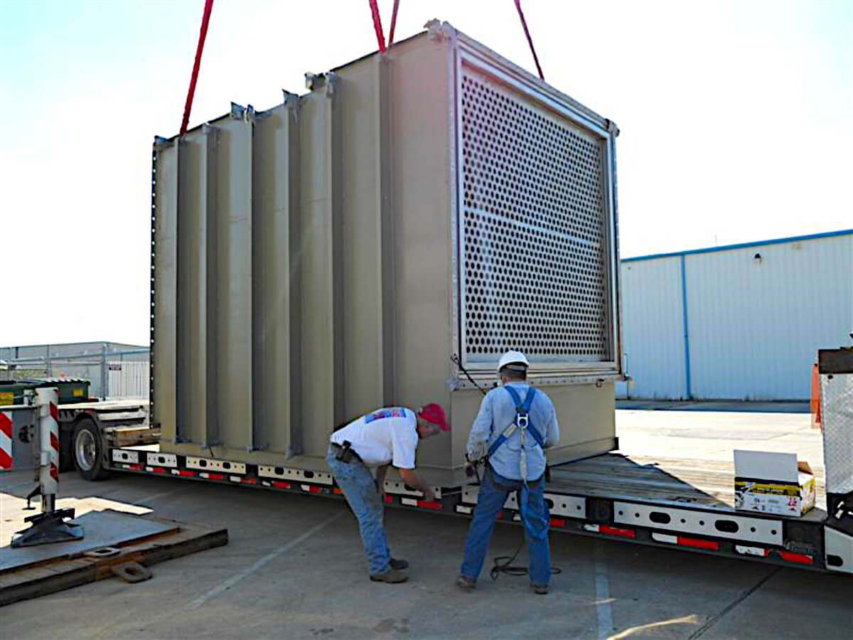 A large square heat exchanger being lowered onto a flatbed trailer bed from a crane. Two workers with hardhats are securing it down