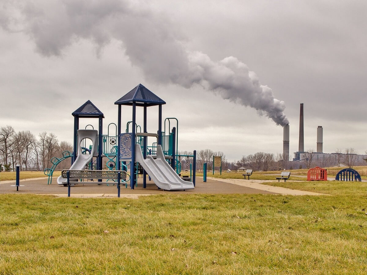 Children’s Colorful Playground Equipment In Park with Green Grass Situated Right Next to Smoking Industrial Smoke Stack Against Grey Sky