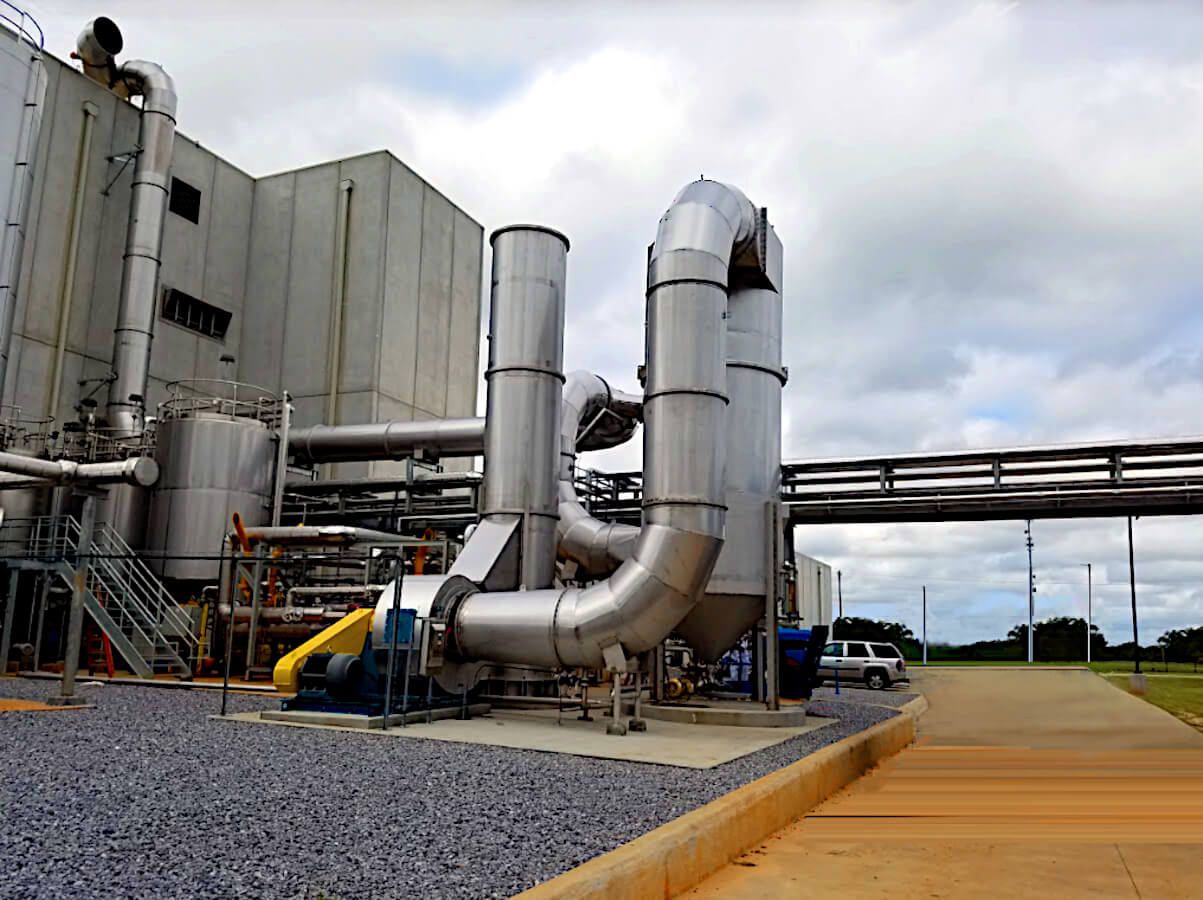 Full View of Silver Stainless Steel Multi-Vane Venturi Scrubber System Abating Particulate Matter (PM) at a Light Grey Food Processing Plant. A Yellow Road Beside the Scrubber Extends Back to a Parking Lot with a Silver Car. The Scrubber Vessel, Stack and the Grey Building Extends Up Into Grey Cloudy Sky with Some Blue Peaking Through.
