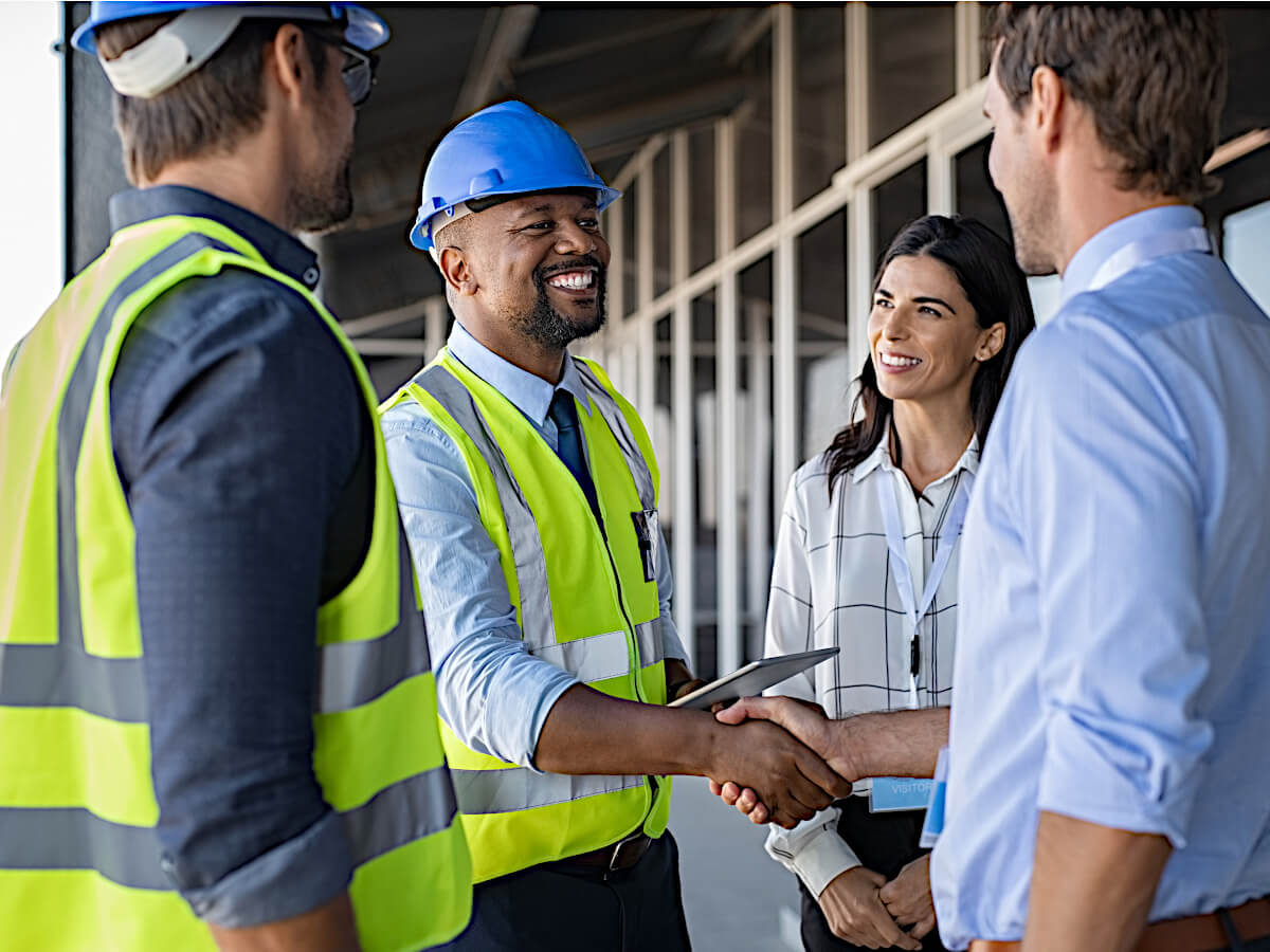 Two Men From Same Company Wearing Yellow Safety Vests and Hard Hats Shake Hands with a Visiting Male and Female Engineering Team