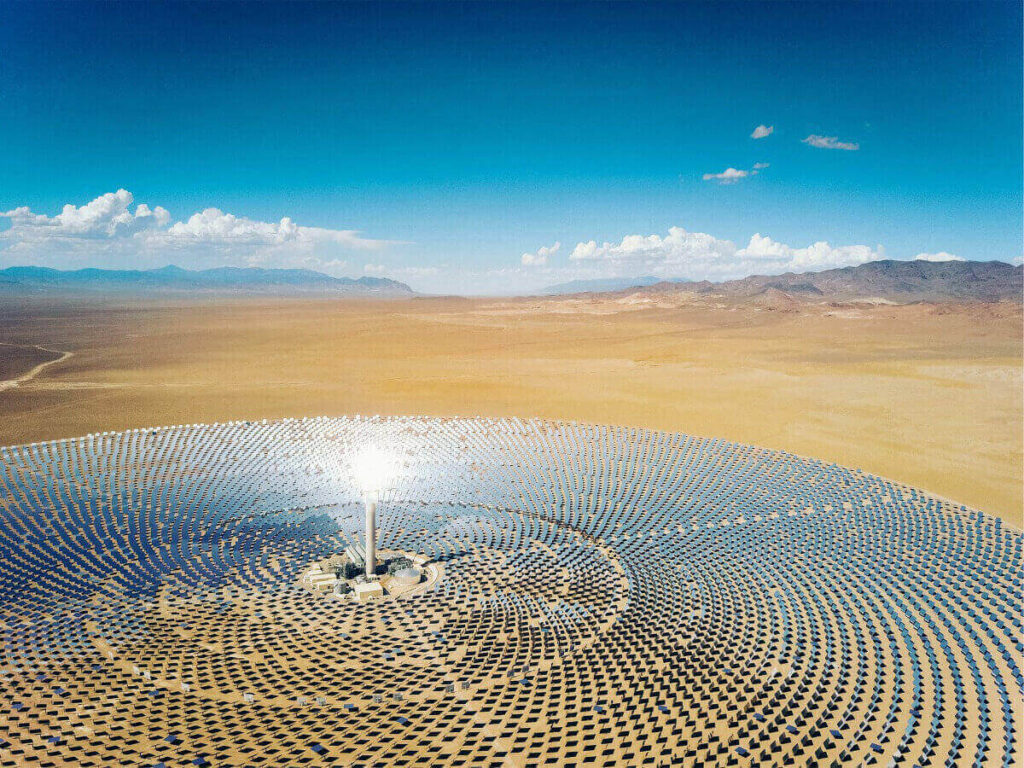 A Vast Array of Silver Solar Panels Surround A Central Control Hub like a BullsEye at a Solar Power Station. Tan Colored Sand and Mountains in the Background contrast with the deep Blue Sky. The Solar Plant Converts Energy From the Sun into Electricity