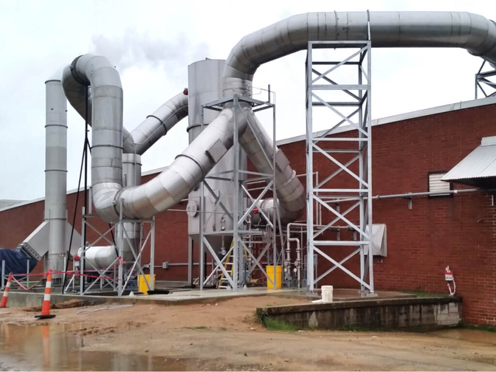 A large Stainless Steel Multi-Vane (MVS) Scrubber is Installed and Waiting to be Commissioned at Building Materials Plant. The Stainless Steel MVS has Branching Ductwork Winding Around the Main Scrubber Vessel and attaches to a Red Brick Building against a White and Blue Sky.