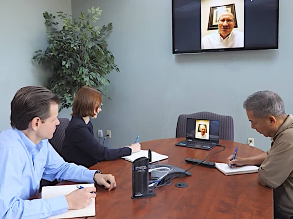 Three Engineering Team Members Sit at a Conference Table and Consult with Potential Customer VIA Zoom on Wall-mounted Computer Screen About their Plant’s Process Application and Emissions Requirements