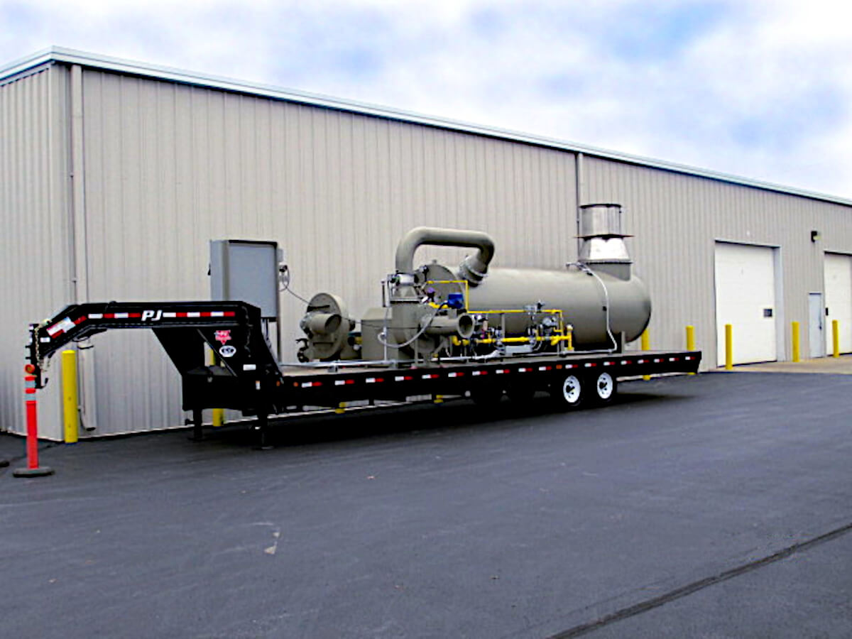 Green Trailer-Mounted Portable Thermal Oxidizer on a Black Trailer parked in front of a tan building against a blue sky with white clouds. It is ready to be transported to a Location for Pipeline Degassing