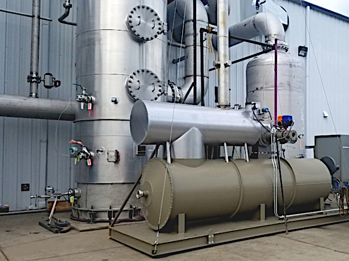 VOCs Destroyed by Green Recuperative Thermal Oxidizer at Chemical Plant. The Recup TO is Connected Via Stainless Steel Ducts to a Tall Stainless Steel Vessel in Front of a Silver Building.