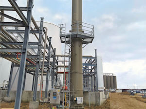 Green Vertical Enclosed Flare Abates VOC at Petrochemical Facility. The Enclosed Flare Stands Before White and Brown Chemical Tanks. Grey Support Structures Hold Piping from the Tanks to the Flare. The Ground is Brown and the Sky is Blue with White and Grey Clouds.