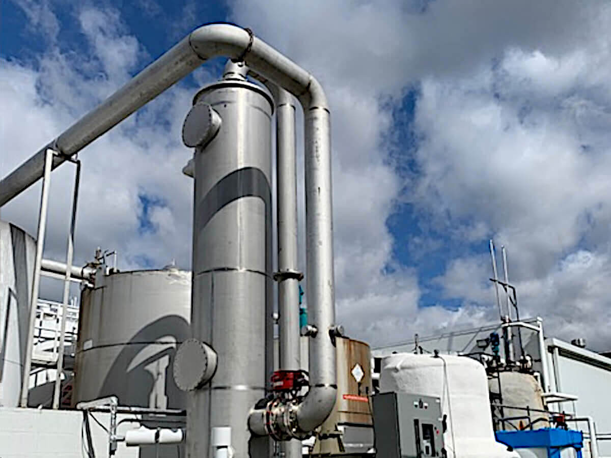 Silver Stainless Steel Wet Scrubber Abates Hydrazine for Chemical Industry. The Scrubber is surrounded by White,, Silver and Rusty Metal Chemical Storage Tanks against a Blue Sky with Grey and White Clouds. It is a Sunny Day.