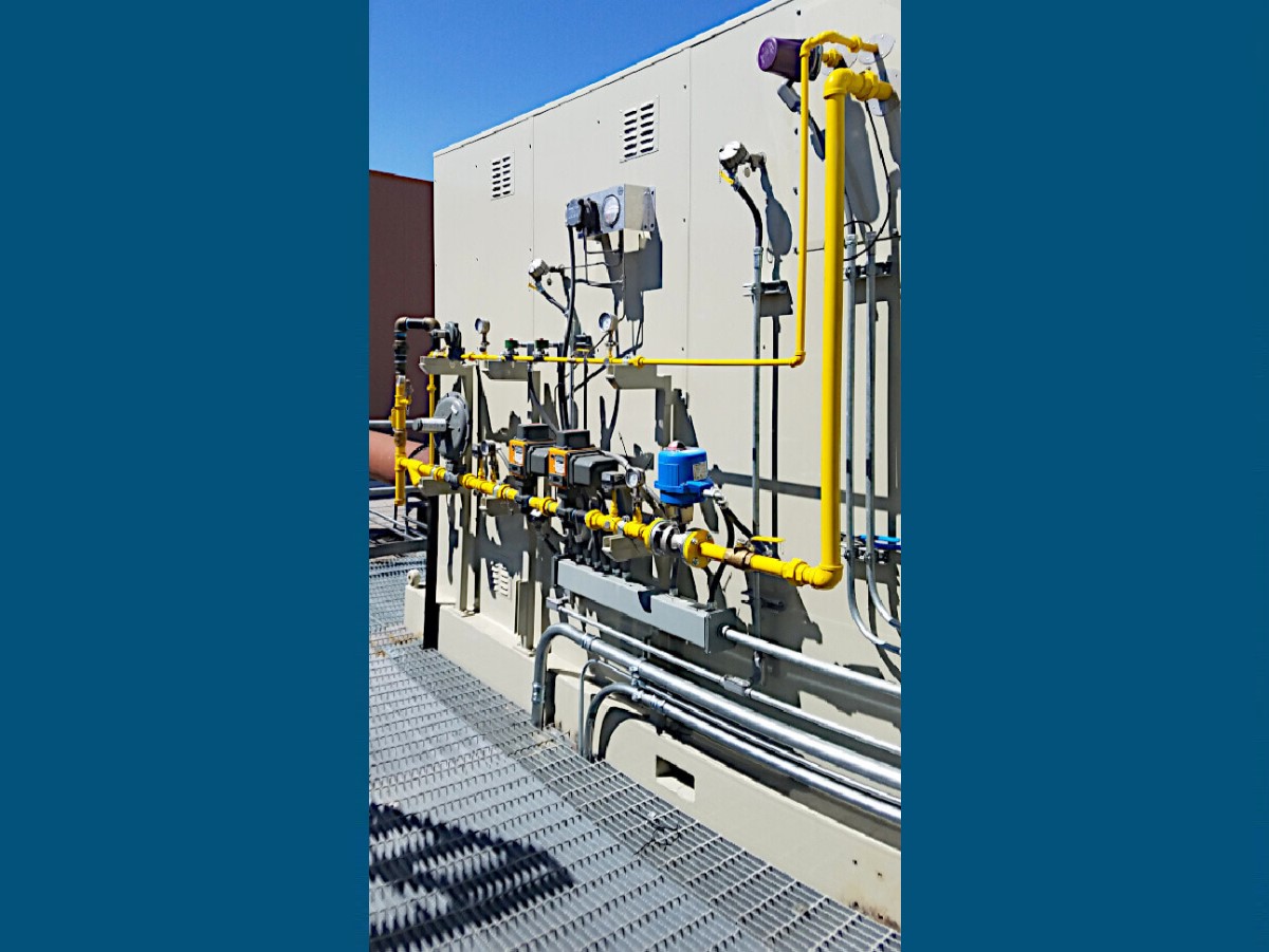Closeup View of Tan Catalytic Oxidizer with Bright Yellow and Silver Piping and Blue Gauges Sits on a Grey Stainless Steel Platform against a Brown Building. It is a Bright Sunny Day with a Clear Blue Sky.
