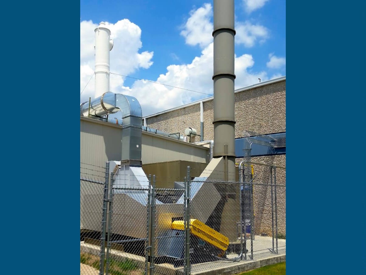 A Dark Green Recuperative Catalytic Oxidizer with a Blue and Yellow Fan and Silver Ductwork Abates all EtO Emissions from a Medical Sterilization Facility. The Oxidizer's Light Green Stack Extends up into a Blue Sky with White Fluffy Clouds on a Bright Sunny Day.