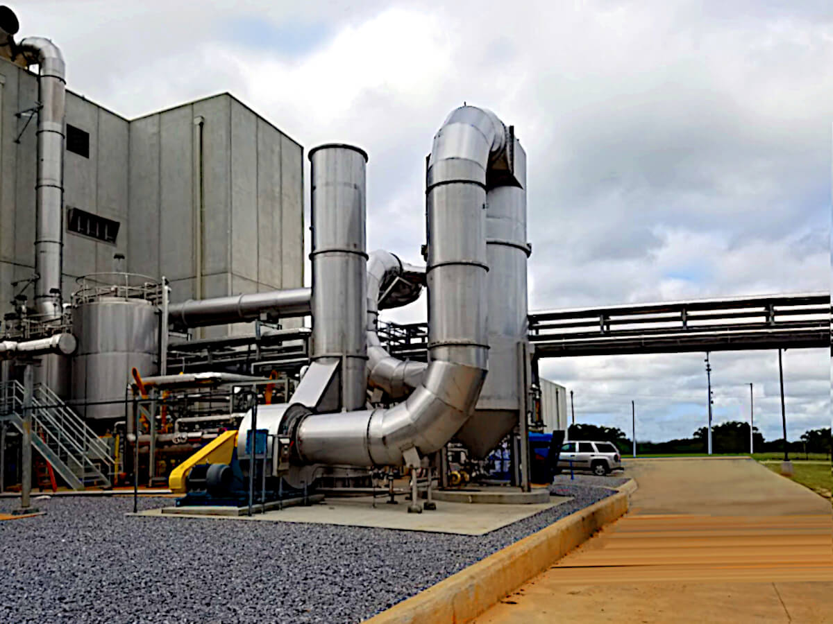 Full View of Silver Stainless Steel Multi-Vane Venturi Scrubber System Capturing PM 2.5 at a Light Grey Food Processing Plant. A Dark Yellow Road Beside the Scrubber Runs Back to a Parking Lot with a Silver SUV. The MVS Scrubber Vessel, Stack, Grey Building and Horizontal Piping are Shown against a Grey Cloudy Sky with Some Blue Peaking Through.