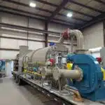 A french gray thermal oxidizer system with a blue fan is ontop of a black trailer inside of a manufacturing warehouse.