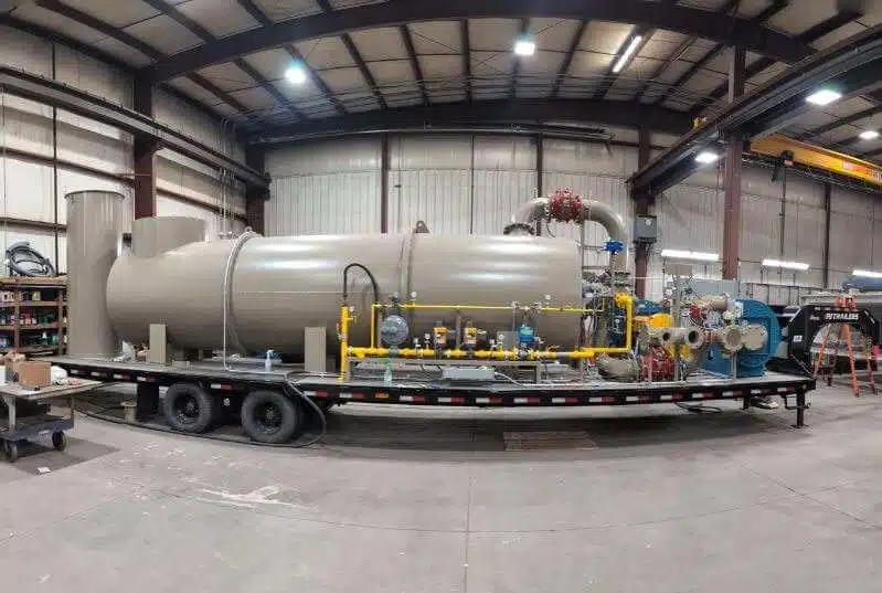 The side view of a french gray thermal oxidizer system, which has a yellow gas train and sits ontop of a black trailer inside of a manufacturing warehouse.