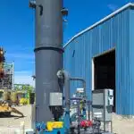 A dark grey enclosed flare standing vertically outside on a concrete pad. It is adjacent from a large blue warehouse, which is visbly behind the enclosed flare on the far right.