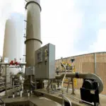 Vertical Enclosed Flare (left-centered) installed outdoors at an idustrial site. The control panel and fan are to the right of the flare.